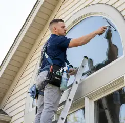 Professional window cleaner on a ladder using a squeegee to clean an exterior arched window, Tuscaloosa, AL.